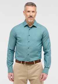 Tee kleurige langmouwshirt met een gestructureerd patroon, knopenlijst aan de voorkant en contrasterende donkere knopen, gecombineerd met beige broek en een bruine riem.