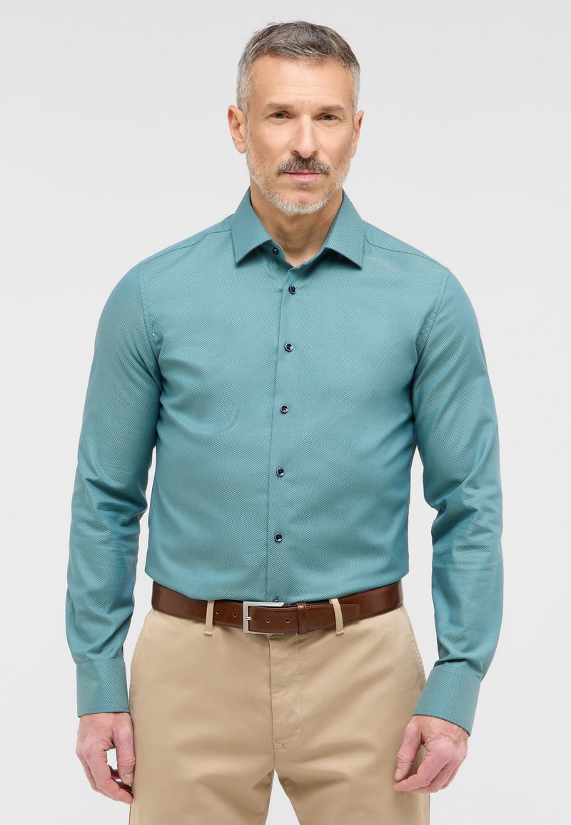 Tee kleurige langmouwshirt met een gestructureerd patroon, knopenlijst aan de voorkant en contrasterende donkere knopen, gecombineerd met beige broek en een bruine riem.