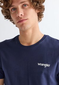 Ung mand med krøllet hår iført en mørkeblå Wrangler t-shirt mod en ensfarvet lys baggrund.