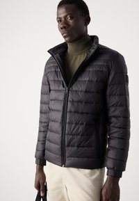 Zwarte geïsoleerde jas met horizontale gestikte patronen, ritsluiting, opstaande kraag en elastische manchetten. Draagt over een donkergroen turtleneck.