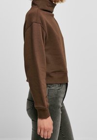 Persona con sudadera de cuello alto marrón y vaqueros negros desgastados mostrada de perfil contra un fondo liso y claro.