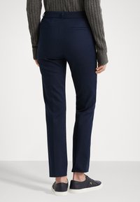Pantaloni navy su misura con vestibilità slim, realizzati in un tessuto morbido, dotati di tasche posteriori e una leggera lucentezza, abbinati a sneakers nere.