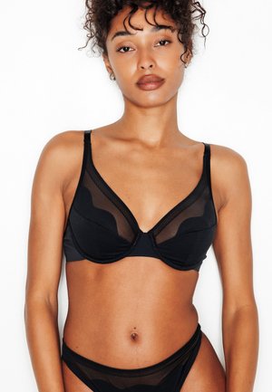 Femme aux cheveux bouclés portant une lingerie noire transparente, debout devant un fond blanc avec une expression neutre.