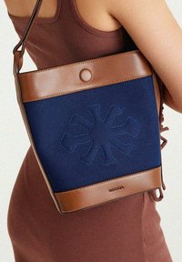 Blue canvas handbag with brown leather accents, featuring a floral embossed design and a circular snap closure. Adjustable strap included.