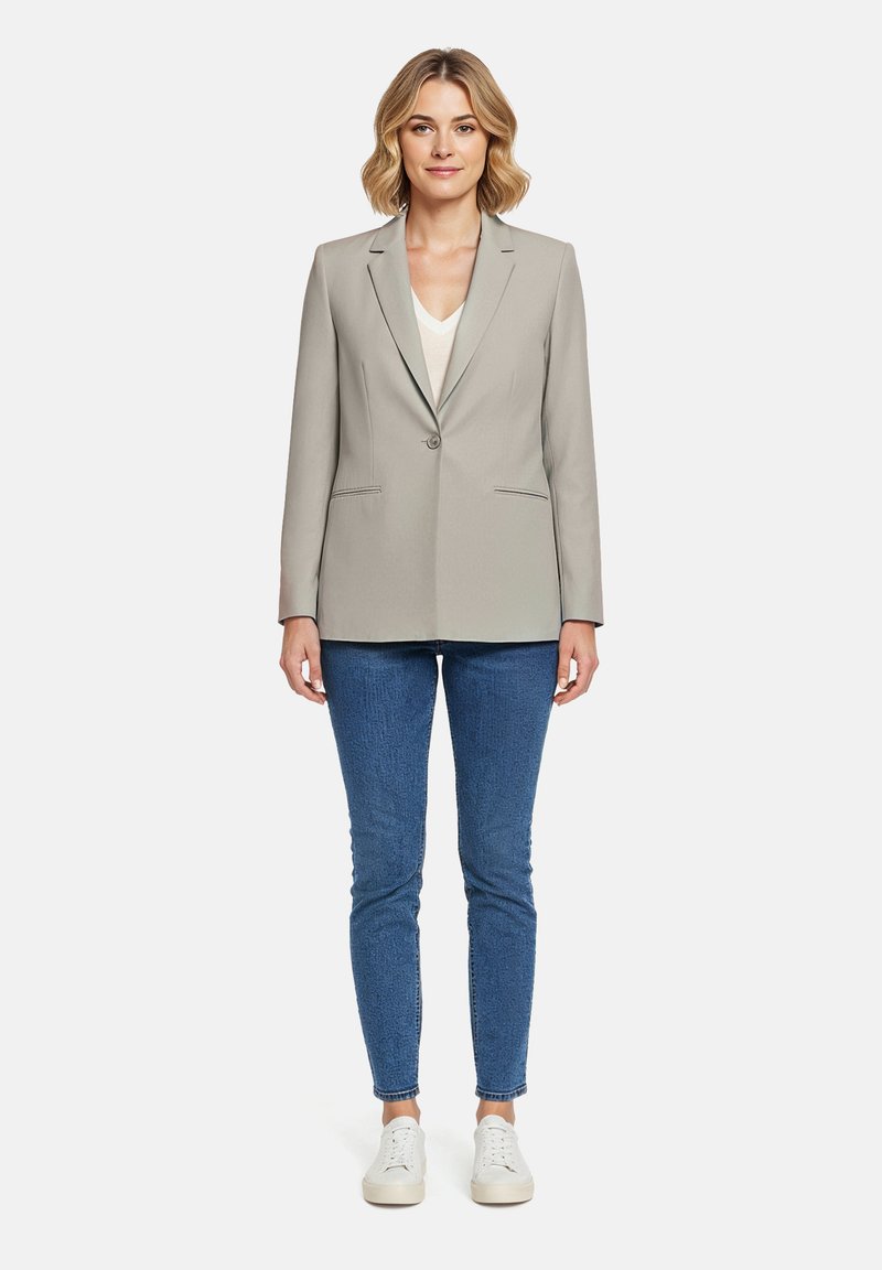 Femme debout, de face, portant un blazer beige, un haut blanc, un jean bleu et des baskets blanches, sur un fond uni.