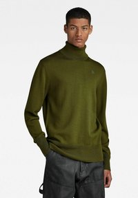 Olivgrüner Rollkragenpullover aus weichem Strickmaterial, mit figurschmeichelndem Schnitt und gerippten Bündchen, kombiniert mit dunklen Hosen.