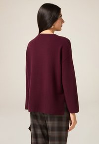 Bordeauxkleurige gebreide trui met een relaxte pasvorm, verlaagde schouders en een geribde zoom. Gecombineerd met bruine geruite broek, met een ingetogen ontwerp.