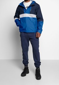Giacca a vento blu e blu scuro con una striscia bianca sul petto, abbinata a pantaloni della tuta blu scuro e stivali neri.