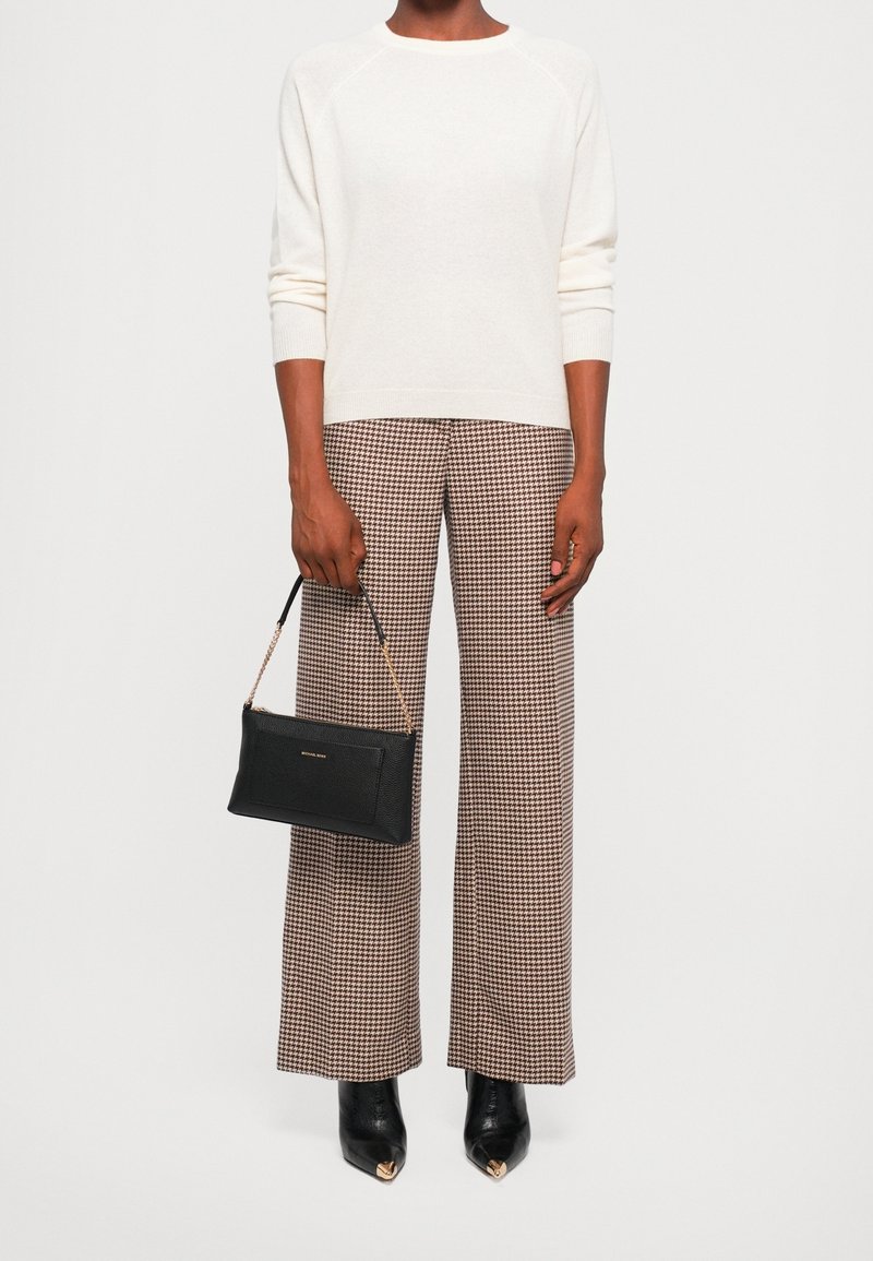 Personne portant un pull blanc, un pantalon large marron à motif pied-de-poule, des bottines noires à bout pointu, tenant un petit sac à main noir avec une bandoulière chaîne dorée.