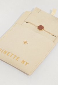 Beige velúr táska arany betűkkel, egy arany lánc karkötővel, amelyen egy kerek, barna kő dísz található. Kattintós zár.
