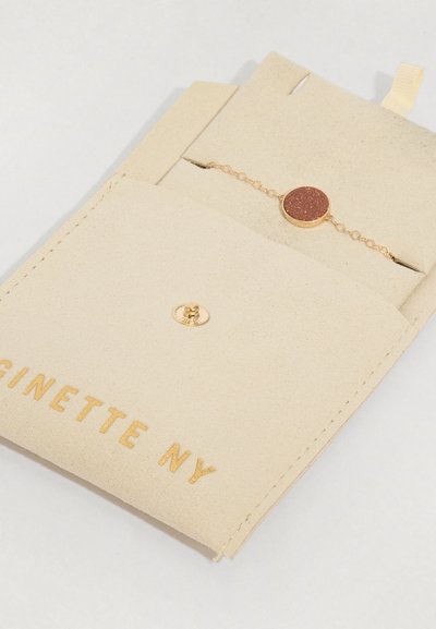 Beige nahkakankainen pussi, jossa on kultakirjontaa, sisältää kultaketjun rannekorun, jossa on pyöreä ruskea kiviaksentti. Kiinnitys napin avulla.