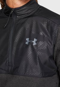 Primer plano de una chaqueta deportiva negra con cremallera y un sutil patrón de puntos, con el logotipo gris de Under Armour en el pecho.
