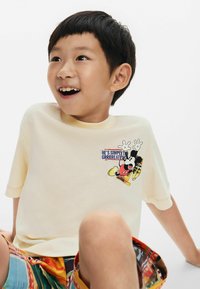 Krem majica s kratkim rukavima s grafikom karikaturice i tekstualnim detaljem na prsima; nosi se s šarenim kratkim hlačama s uzorkom.