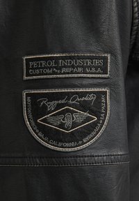 Zwarte leren jas met een getextureerde afwerking. Twee patches: "Petrol Industries" en een ronde afbeelding met vleugels en "Rugged Quality".