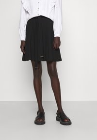 Zwarte geplooide rok met een tailleband die je kunt strikken, gecombineerd met een witte blouse met lange mouwen en ruches. Zwarte sandalen met open tenen maken de look compleet.