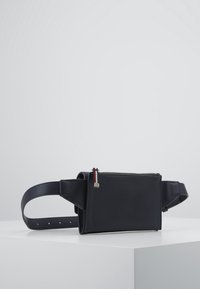 Černá kožená taška přes pás s texturovaným povrchem, obdélníkovým tvarem, zipovým uzavíráním a malou dekorativní visačkou s tříbarevným akcentem.