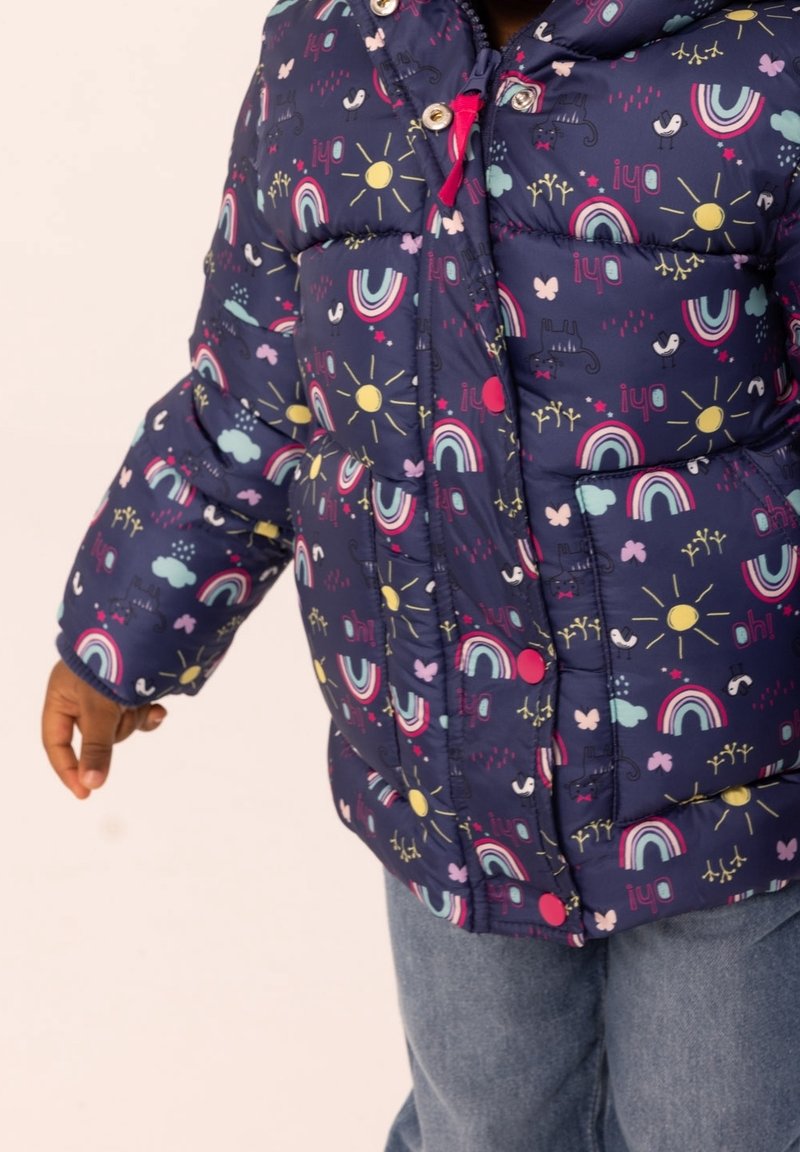 Enfant portant une veste matelassée bleu marine avec des arcs-en-ciel colorés, des soleils, des nuages, des oiseaux et des papillons, assortie à un jean bleu.