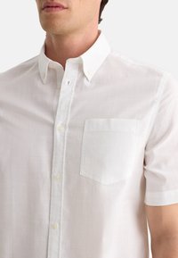 Camisa blanca de manga corta en tela ligera, con un cuello tipo button-down y un bolsillo en el pecho izquierdo con un diseño cuadrado.