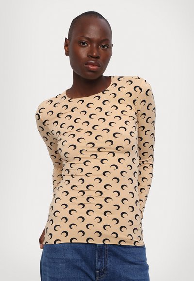 Top de manga larga, ajustado, en tela beige con un patrón de luna creciente negra. Cuello redondo y textura suave. Combinado con jeans azules.