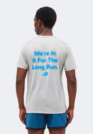 Mann, der mit dem Rücken zur Kamera steht, trägt ein graues Shirt mit blauem Schriftzug „We're In It For The Long Run“ und blaue Sportsshorts vor einfachem Hintergrund.