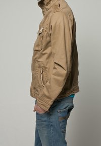 Beżowa kurtka na zamek z wysokim kołnierzem, bocznymi kieszeniami i kieszenią na piersi. Nosi się ją na niebieską koszulę, w połączeniu z niebieskimi dżinsami.