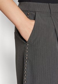 Main dans la poche d’un pantalon gris à fines rayures, avec des rangées verticales de clous argentés brillants le long de la couture latérale.