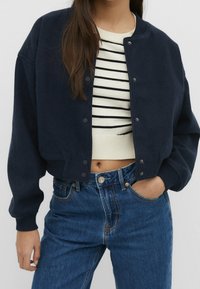 Marineblauwe cropped bomberjack met een geribbelde onderrand, drukknopen en verlaagde schouderzomen, gedragen over een gestreepte trui en hoge taille jeans.