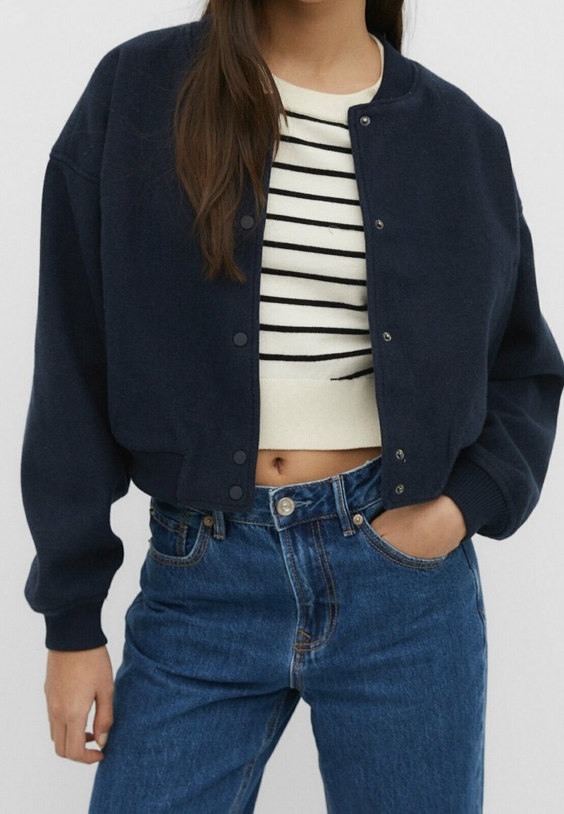Marineblauwe cropped bomberjack met een geribbelde onderrand, drukknopen en verlaagde schouderzomen, gedragen over een gestreepte trui en hoge taille jeans.