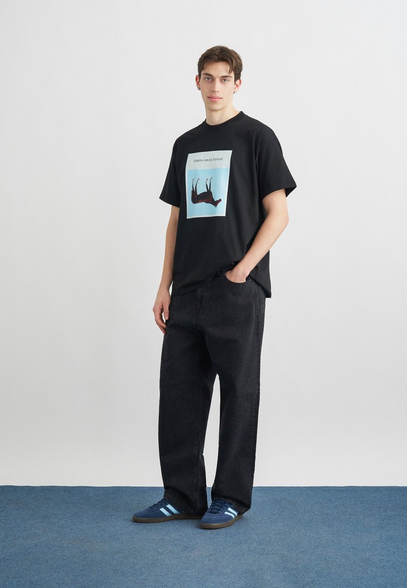 Junger Mann steht, trägt ein schwarzes Grafik-T-Shirt mit aufgedrucktem, auf dem Kopf stehendem Tiermotiv, lockere schwarze Hose und blaue Turnschuhe auf blauem Teppich.