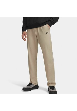 Uomo che indossa pantaloni della tuta beige Under Armour, scarpe da ginnastica nere e grigie, e una maglia a maniche lunghe nera, in piedi con una mano in tasca.