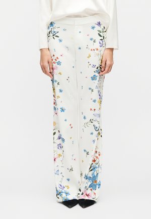 Fehér, bőszárú nadrág, több színű virág- és pillangómintával, fehér hosszú ujjú felsővel és fekete hegyes orrú cipővel viselve.