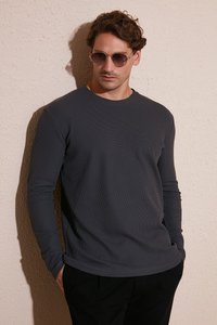 Cămașă lungă de culoare gri închis, cu un design texturat și nervurat. Modelul poartă ochelari de soare rotunzi, stând în fața unui perete bej texturat.