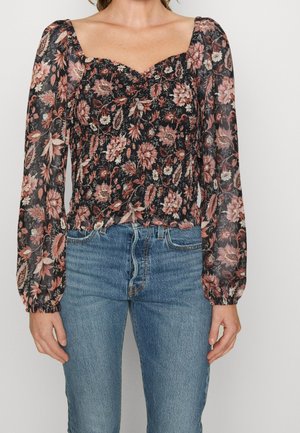 Kobieta nosząca czarną przezroczystą, kwiatową bluzkę z długimi bufkami na rękawach, w połączeniu z niebieskimi dżinsami.