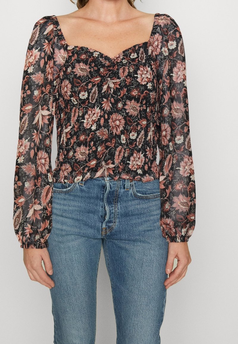 Femme portant un chemisier noir transparent à fleurs à manches longues bouffantes, associé à un jean en denim bleu.