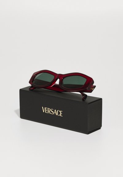 Raudoni kampuoti saulės akiniai su tamsiai žaliais lęšiais, padėti ant juodos dėžutės su auksiniu "VERSACE"logotipu. Lygus plastikinis paviršius, be raštų.