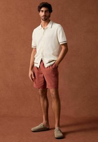 Korte mouw ribgebreid shirt in crème met grijze accenten, gecombineerd met roestkleurige shorts en beige instappers. Bruine achtergrond.
