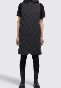 Person wearing a black quilted sleeveless hooded vest over a black short-sleeve shirt, black leggings, and black ankle boots.