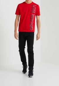 Červené bavlněné tričko s vertikálním černým textem "LOVE MOSCHINO", kombinované s černými džínami a černými sportovními botami.