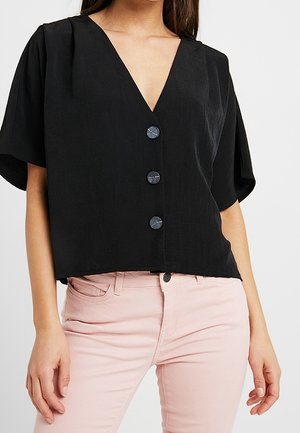 Kvinde iført en sort kortærmet V-hals bluse med tre knapper foran kombineret med lyserøde bukser, stående mod en ensfarvet baggrund.