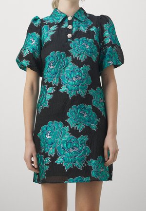 Kobieta ubrana w krótką czarną sukienkę z dużym turkusowym kwiatowym wzorem, bufkami na rękawach i trzema ozdobnymi guzikami na kołnierzyku.