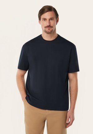 Mann mit kurzen braunen Haaren und Schnurrbart, trägt ein schwarzes kurzärmeliges Hemd und beige Hose, steht mit einer Hand in der Hosentasche, neutraler Hintergrund.