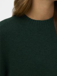 Lähedalt pilt inimesest, kellel on seljas tume roheline kudum, millel on soonikuga ümar kaelus, ja pikk pruun juukseosa on osaliselt nähtav.