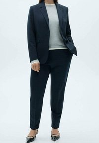 Costume marine avec un blazer tailleur et un pantalon assorti, associé à un haut gris clair. Tissu lisse avec un design structuré et des talons pointus.