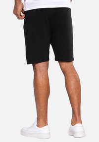 Homme portant un short noir à côtes, des baskets blanches et une chemise blanche, montré de la taille aux pieds, debout sur un fond uni.