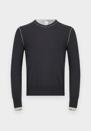 Mladić s kovrčavom kosom stoji s rukama iza leđa, nosi crnu košulju dugih rukava, široke plave traperice i crne cipele.