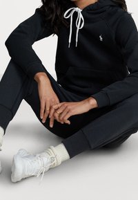 Svart bomullshoodie med vit dragsko och liten logotyp, tillsammans med matchande joggers och vita sportskor med ribbade strumpor.