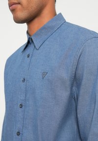 Modrá chambray košeľa s golierom na gombíky, štruktúrovaný strih, s trojuholníkovým logom na ľavej hrudi a jemne textúrovaným povrchom.