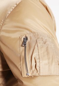 Chaqueta de color beige claro con una textura suave y brillante. Cuenta con un bolsillo con cremallera en la manga izquierda y detalles cosidos.