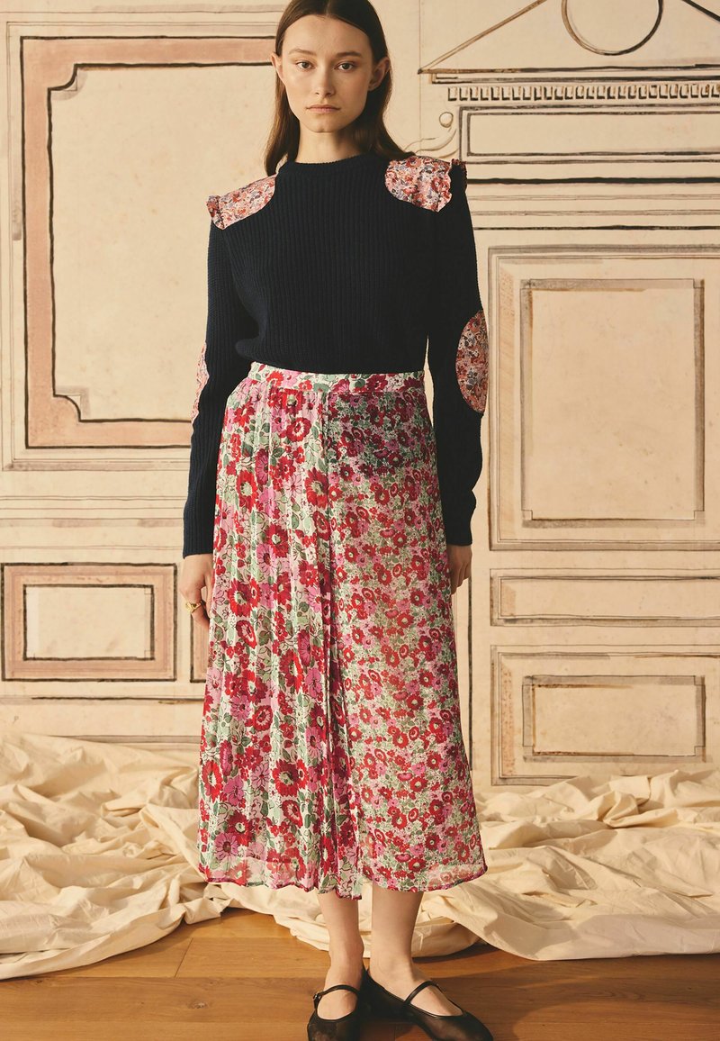 Pull en maille bleu marine avec des accents de patch floraux sur les épaules, associé à une jupe plissée à motif floral dans des tons de rose et de vert.