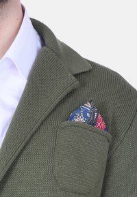 Cardigan in maglia verde oliva con colletto a scollatura, dotato di una tasca sul petto che tiene un fazzoletto a fantasia colorata.